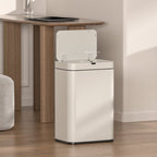 A modern white automatic open-lid canister trash bin placed next to a wooden desk in a bright, minimalist room with light wood floors.