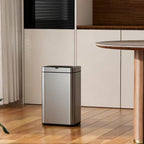 A modern stainless steel automatic floor trash can with a brushed metal finish, set on a wooden floor in a contemporary kitchen environment next to a wooden dining table.