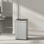 A modern silver and black automatic floor trash can with a touchless lid, set in a minimalist room with a white wall, gray marble floor, and a black grand piano in the background.