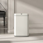 A tall, minimalist white floor-standing appliance with a rectangular body and rounded top, placed in a bright interior room with a piano, lamp, and open book visible in the background.