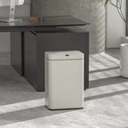 A modern white rectangular smart trash can with a touchless lid stands on a gray concrete floor in a minimalist office setting next to a dark desk and a potted plant.