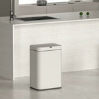 A modern white rectangular trash can with a lid, placed on a light gray floor in a minimalist kitchen setting with a white island and wood stool.